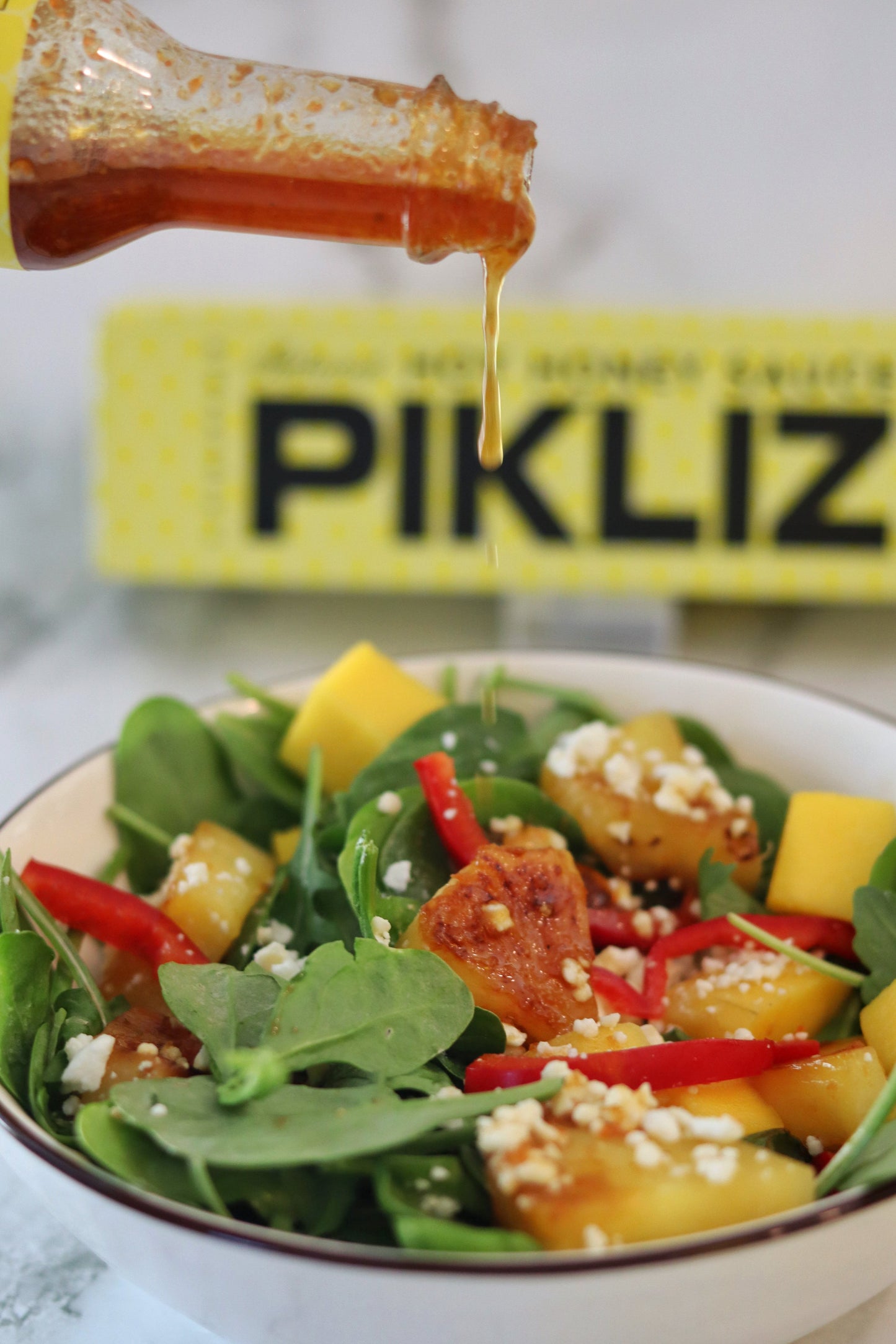 Alexandra’s Pikliz® Hot Honey Sauce pouring from a 6 oz bottle over a tropical pineapple and mango salad