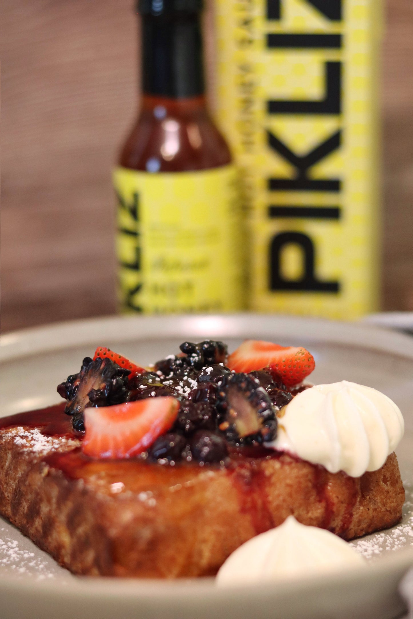 French Toast topped with hot honey caramel sauce made using Alexandra’s Pikliz® Hot Honey Sauce. The plate is styled with berries, and the Alexandra’s Pikliz® Hot Honey Sauce bottle with designer box is blurred in the background.